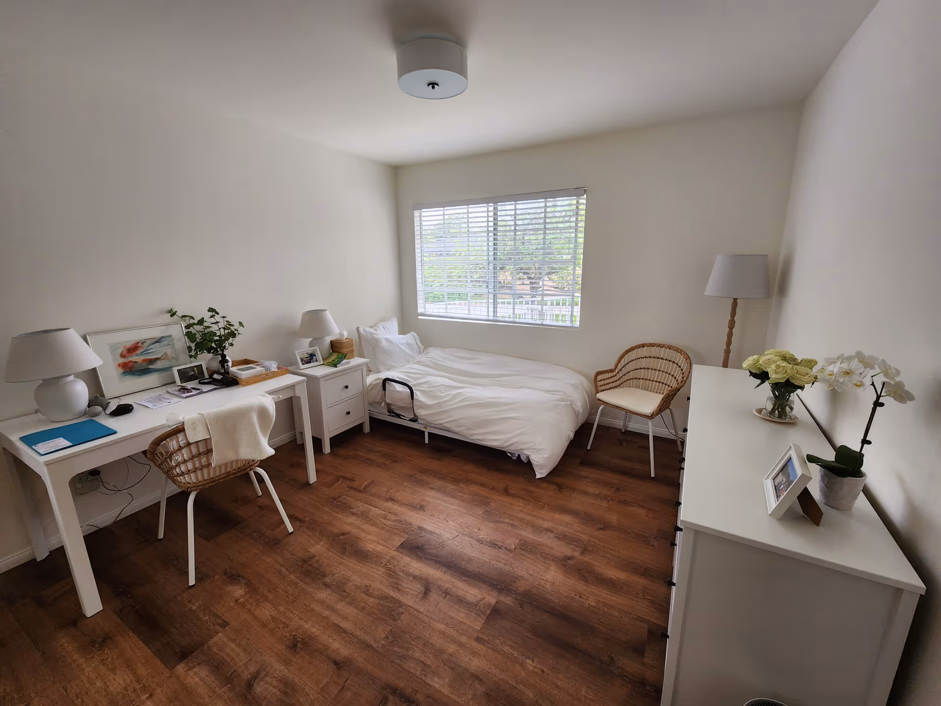 Bright, simply furnished bedroom with a single bed beneath a window, desks, wicker chairs, and a white dresser.
