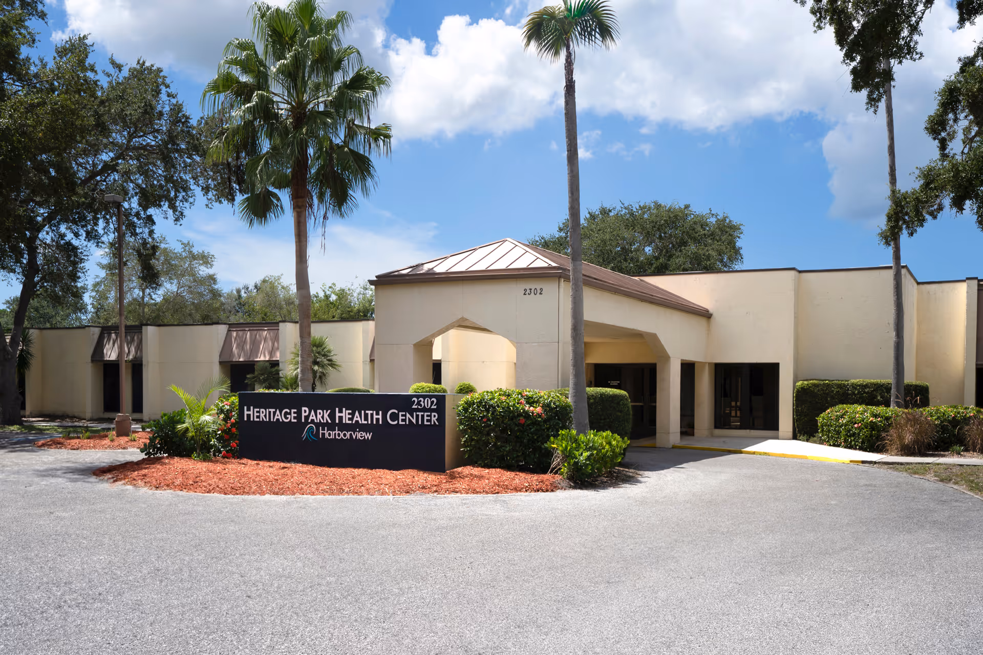 Front exterior of Heritage Park Health Center with an entrance canopy, landscaped sign, driveway, and palm trees under a blue sky.