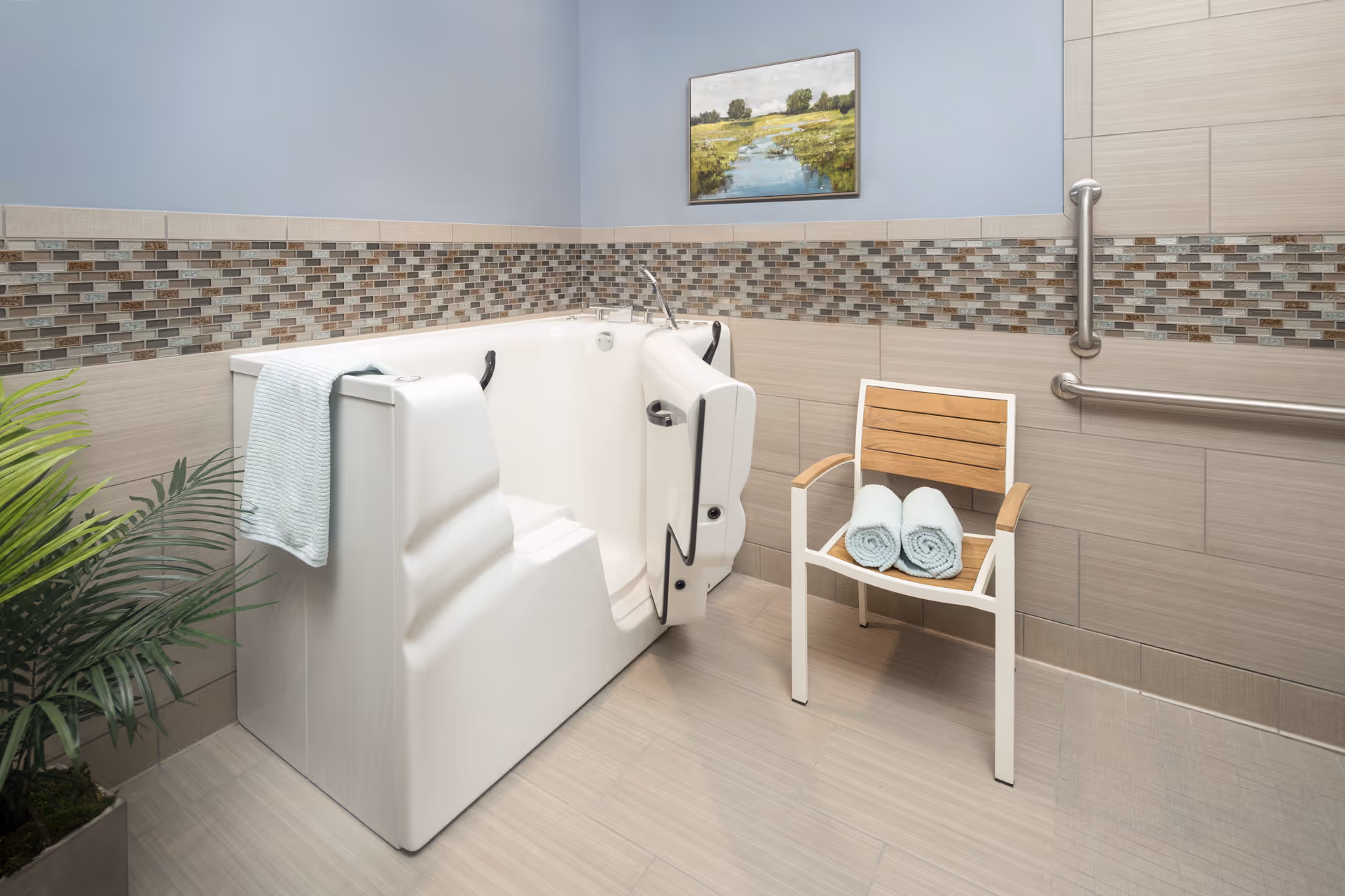 A bathroom with a walk-in bathtub featuring a door and handrails, a wooden chair with two rolled towels on it, a potted plant, and a landscape painting on the wall. The walls have a decorative tile border and grab bars for safety.
