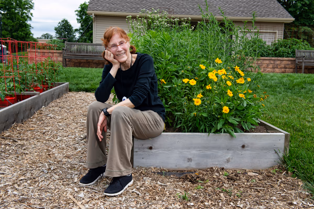 An elderly woman with red hair and glasses sits on the edge of a raised garden bed filled with yellow flowers. She is smiling and resting her head on her hand. The garden area has wood chip ground cover, green grass, and wooden benches in the background near a building.