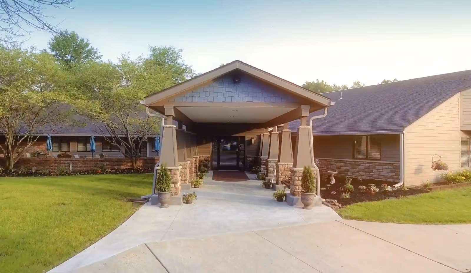 Front entrance of a single-story building with a covered porte-cochère, columns, potted plants, and surrounding lawn.