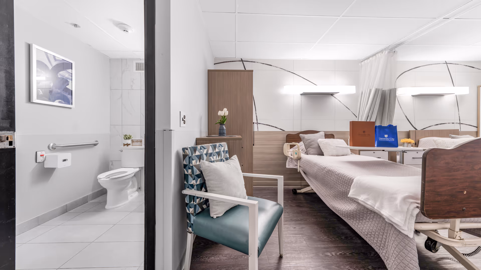 A clean and modern hospital-style room with a single bed covered in a light gray quilt, a padded chair with a geometric pattern cushion, a wooden cabinet, and a small table with bags on it. To the left, there is an accessible bathroom with a toilet, grab bars, and a framed picture on the wall.
