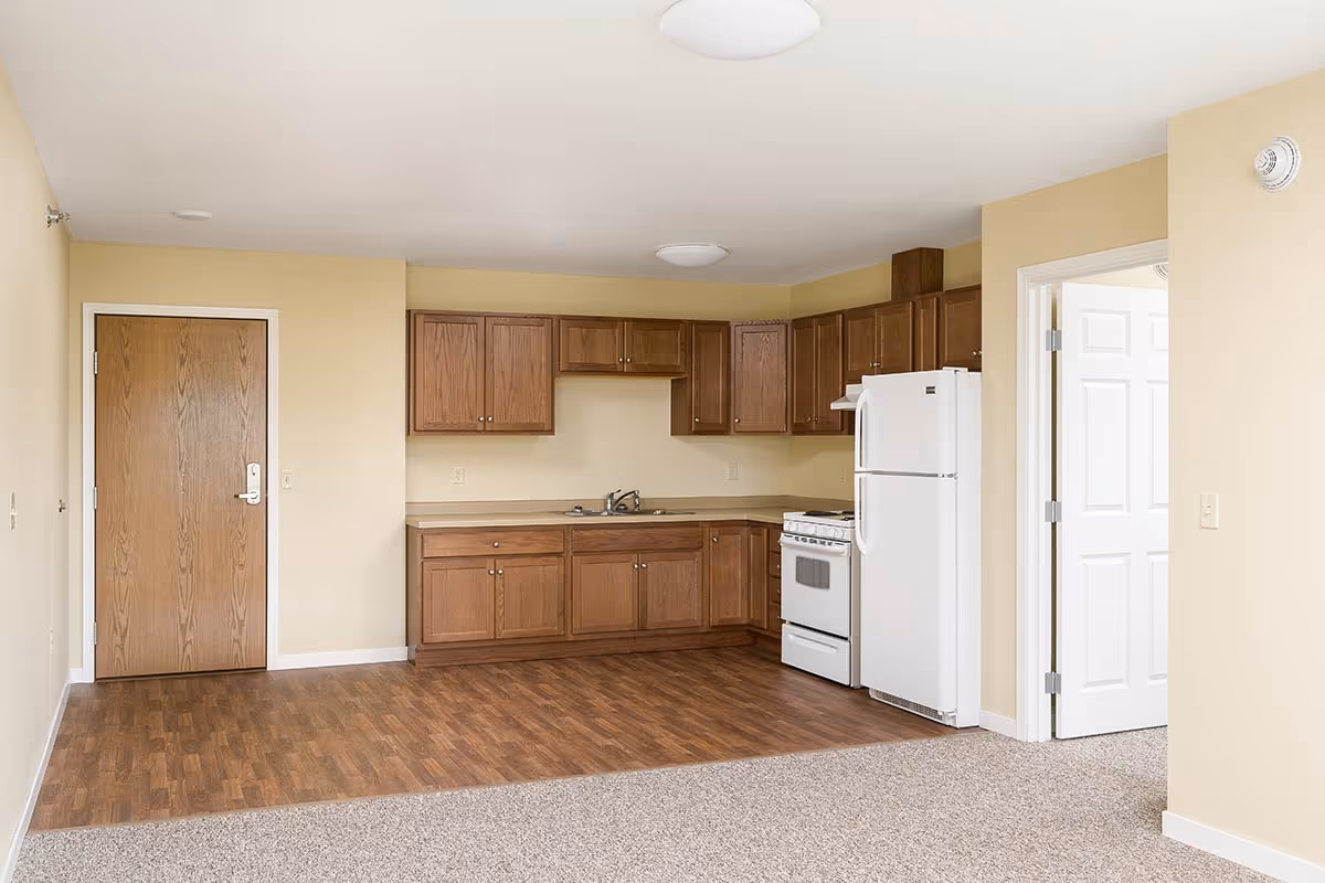 Interior view of a senior living apartment kitchen area with wooden cabinets, a white refrigerator, a white stove, and a sink. The floor has a wood finish in the kitchen area and carpet in the adjacent living space. There is a wooden door on the left and a white door on the right leading to another room.
