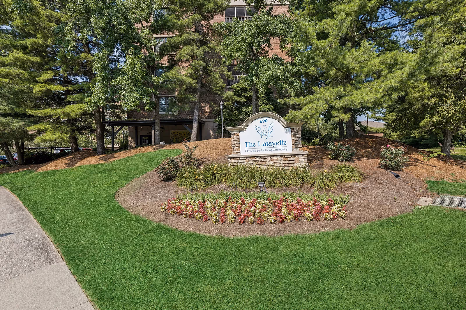 Outdoor view of The Lafayette senior living community sign surrounded by landscaped greenery, flowers, and trees with a building partially visible in the background.