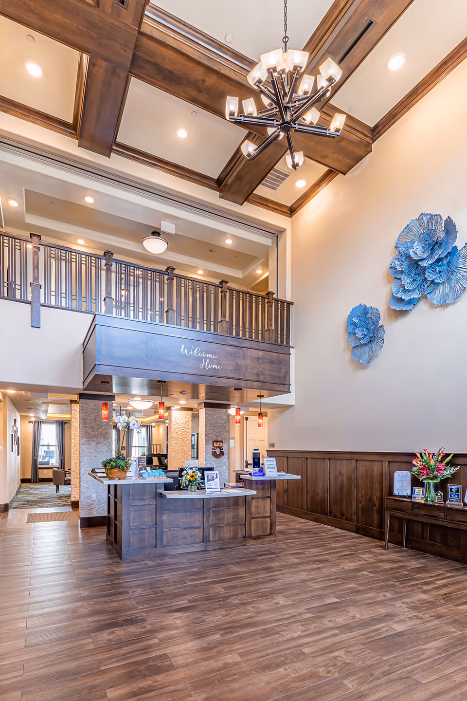 Spacious senior living lobby with a wooden reception desk, high coffered ceiling and chandelier, balcony railing, and blue wall sculptures.