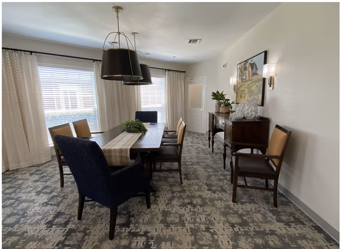 A dining room with a rectangular wooden table surrounded by six chairs, two of which are upholstered in dark blue fabric and the others in tan. The table has a beige runner and a green plant centerpiece. Two large black pendant lights hang above the table. The room has patterned carpet, two large windows with white curtains, a wooden sideboard with decorative items and plants, and a framed painting on the wall.