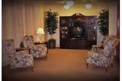 A cozy living room with four floral upholstered armchairs arranged around a wooden entertainment center holding a television. There are two potted plants flanking the entertainment center and a table lamp on a side table next to one of the chairs. The room has beige carpeting and soft lighting from ceiling fixtures.