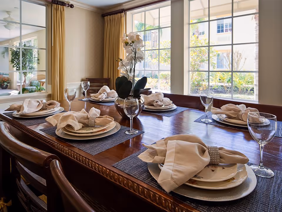 Sunlit formal dining room with a wooden table set with plates, folded napkins, wine glasses, and an orchid centerpiece in front of large windows.