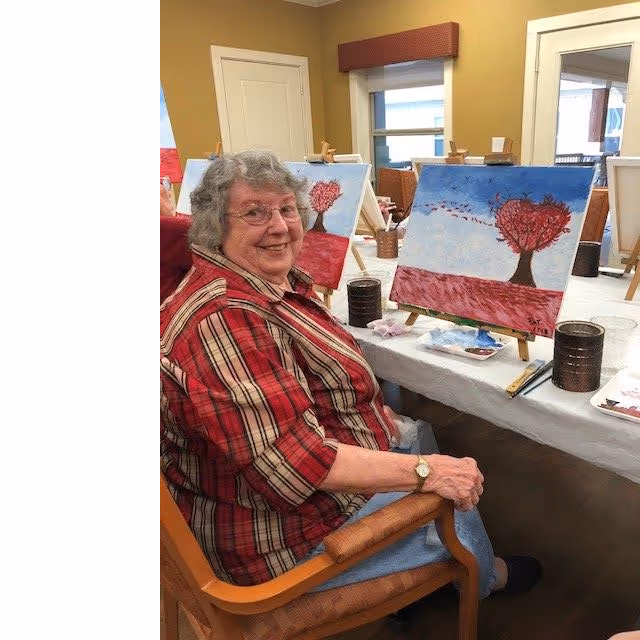 An elderly woman with gray hair and glasses, wearing a red plaid shirt, is sitting on a wooden chair at a table with painting supplies. She is smiling and posing beside her painting of a tree with red leaves shaped like a heart against a blue sky and red ground. The room has beige walls, a window with a red valance, and other easels with similar paintings in the background.