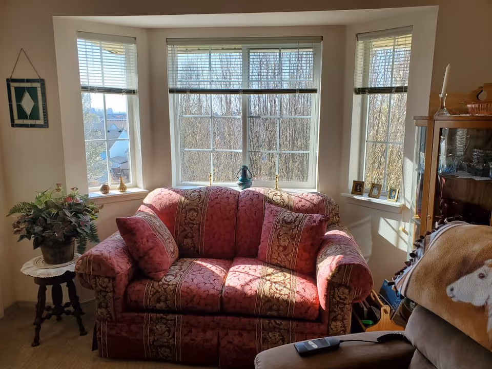 A cozy living room corner with a red patterned loveseat placed in front of three large windows with white blinds. There is a small wooden side table with a potted plant on the left, and a wooden cabinet with glass doors on the right. The room is warmly lit by natural sunlight coming through the windows.