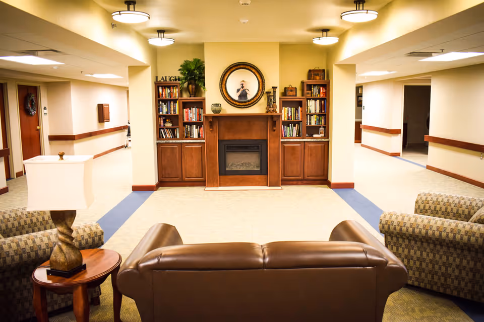 A cozy common area in a senior living facility featuring a brown leather sofa and two patterned armchairs arranged around a wooden side table with a lamp. The background shows a wooden fireplace mantel flanked by built-in bookshelves filled with books and decorative items. Above the fireplace is a round mirror reflecting a person taking the photo. The walls are painted light yellow, and the ceiling has round light fixtures. Hallways extend on both sides of the room.