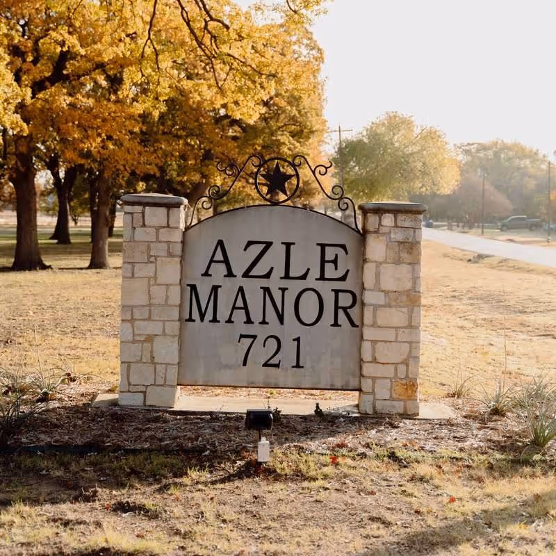 Stone sign for Azle Manor with the number 721, set outdoors with trees and a road in the background during autumn.