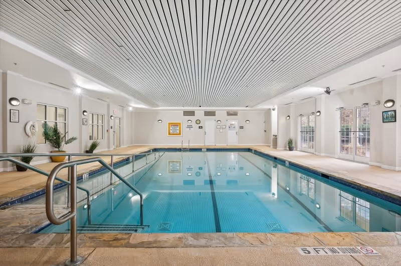 Indoor swimming pool with clear water, metal handrails, and tiled edges. The room has a white ceiling with linear patterns, beige walls, potted plants, and large windows letting in natural light.