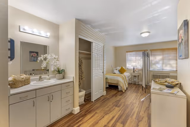 A bright and clean senior living bedroom with wood flooring, a single bed with yellow and white bedding, a small nightstand with a lamp, a closet with sliding doors, a dresser, and a sink with a mirror and countertop on the left side. The room has beige walls and a window with blinds letting in natural light.