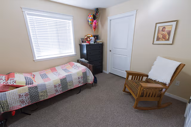 A small bedroom with a single bed covered by a patchwork quilt, a black dresser with balloons and framed photos on top, a wooden rocking chair with a white throw draped over it, a closed white door, a window with white blinds, and a framed picture on the beige wall.