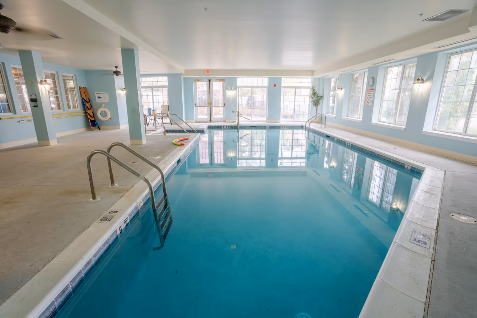 Indoor swimming pool area with clear blue water, metal handrails for entry, large windows letting in natural light, light blue walls, ceiling fans, and pool safety equipment on the wall.