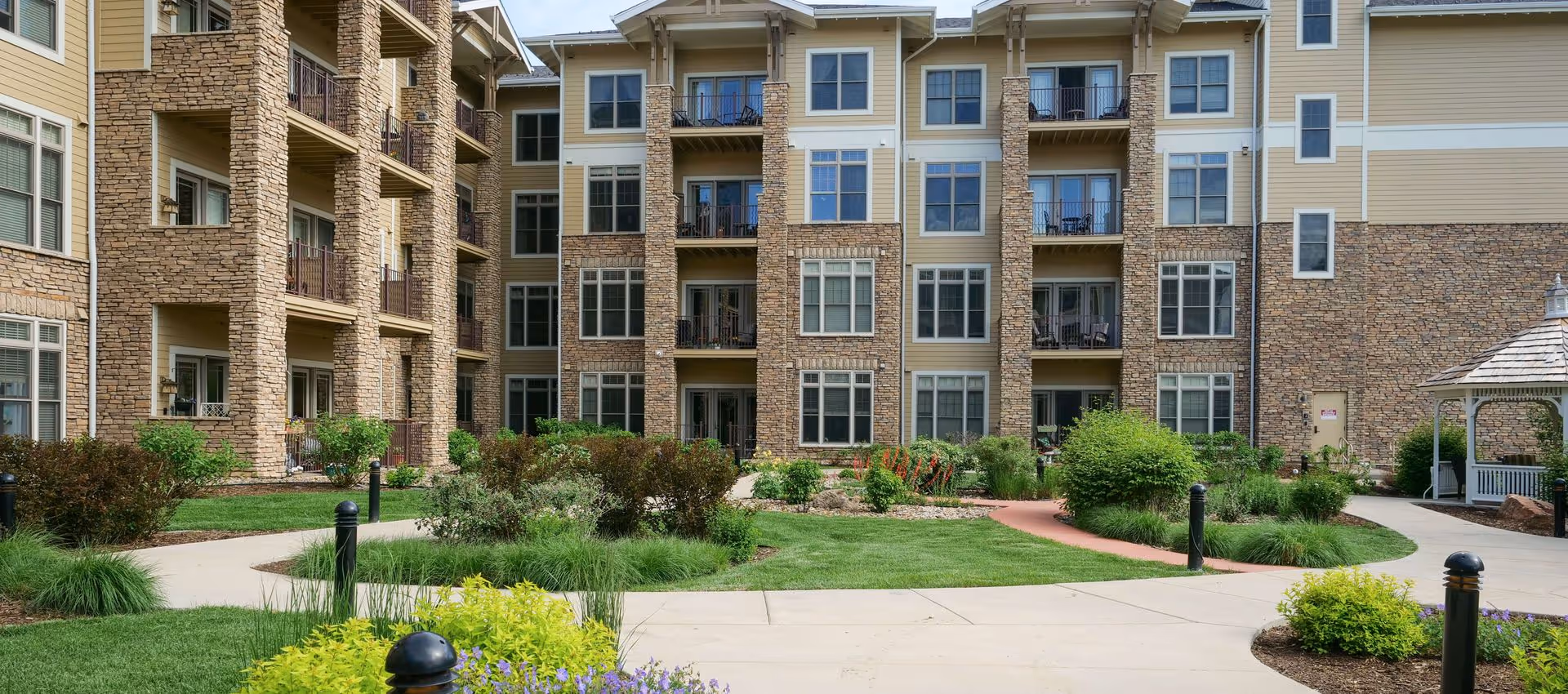 Landscaped courtyard and walkways in front of a multi-story apartment building with stone facade and balconies.