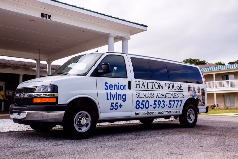 A white Hatton House senior living shuttle van parked under the entrance canopy of a senior apartment building.