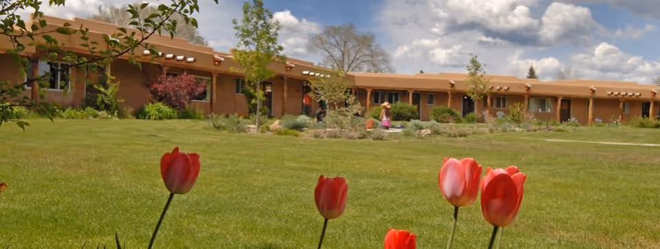 A single-story building with a brown exterior surrounded by a green lawn and garden. Red tulips are in the foreground, and a person is visible near the building. The sky is partly cloudy.