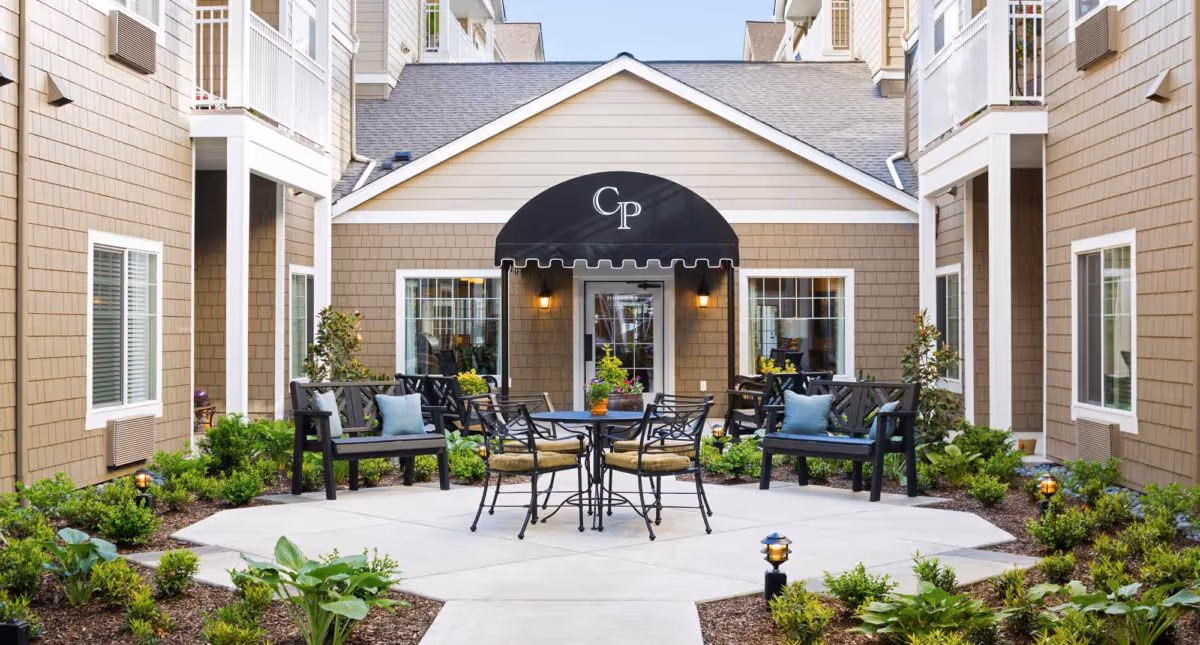 Outdoor courtyard with a round table, chairs and benches in front of a beige building entrance topped by a black 'CP' awning.