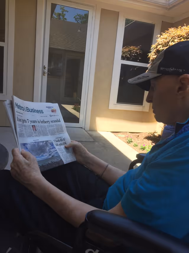 An elderly man wearing a cap and blue shirt is sitting in a wheelchair outdoors, reading a newspaper titled Metro & Business. Behind him is a beige building with a glass door and windows, and some plants in a small garden bed.