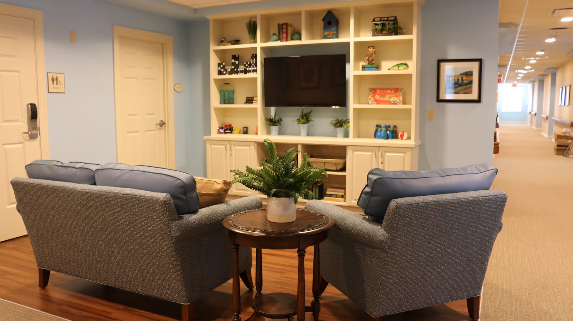 A cozy seating area in a senior living facility with two gray armchairs facing a built-in white shelving unit that holds a flat-screen TV, books, and decorative items. A small round wooden table with a potted plant is placed between the chairs. The walls are painted light blue, and a hallway extends to the right with framed artwork on the walls.