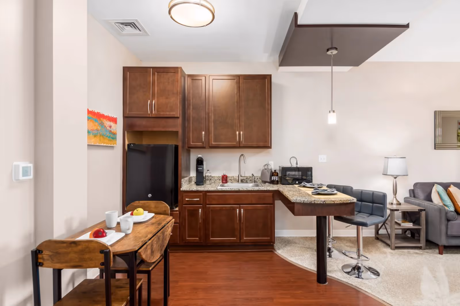 A small kitchen area with dark wooden cabinets, a granite countertop, a black mini refrigerator, a coffee maker, and a microwave. There is a wooden table with two chairs and place settings including cups and plates with fruit. Adjacent to the kitchen is a living area with a gray sofa, a side table with a lamp, and a wall mirror.