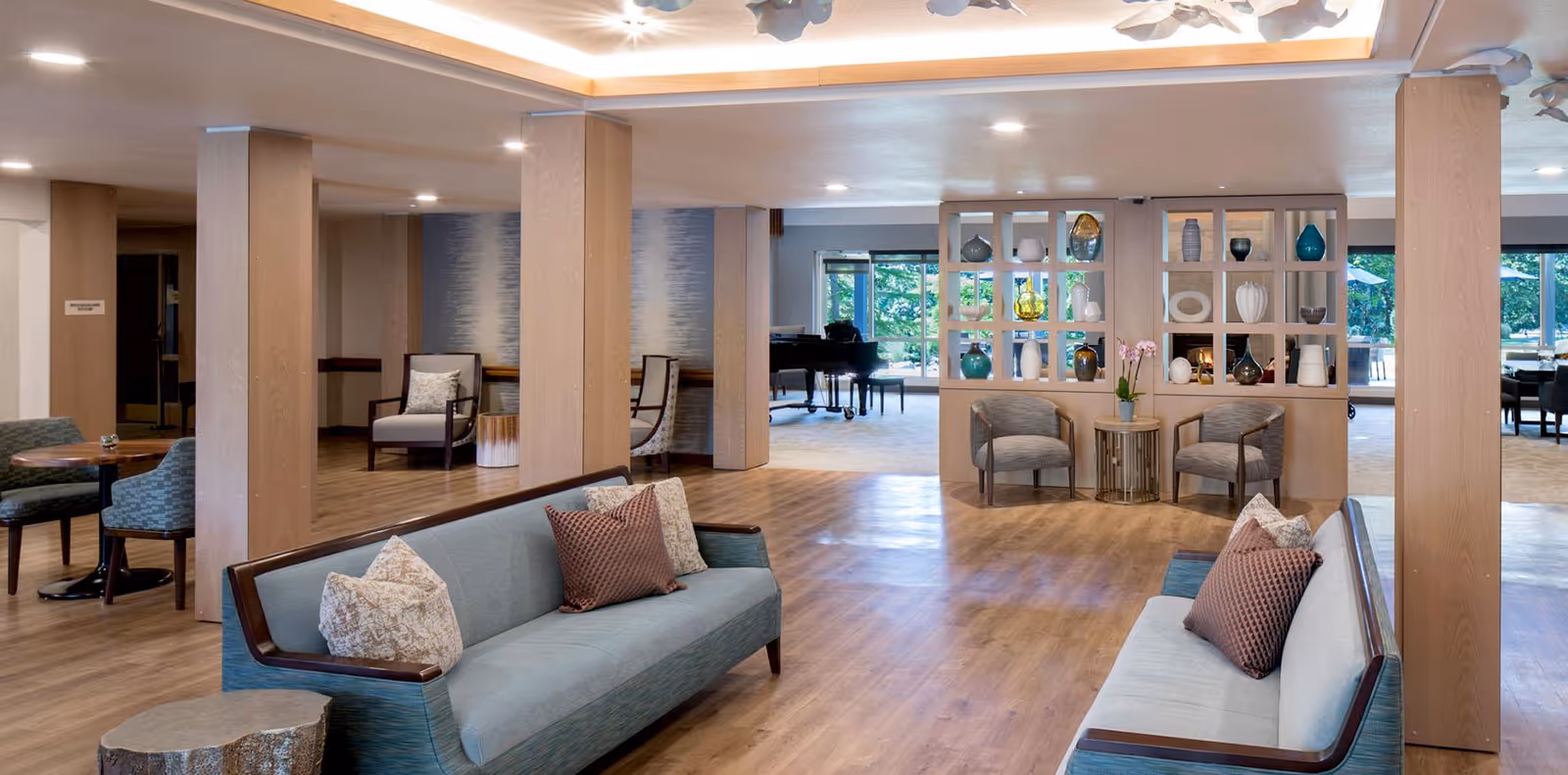 A spacious and well-lit living room area in a senior living facility with two light blue sofas facing each other, each adorned with decorative pillows. The room features wooden flooring, wooden columns, and a decorative partition with various vases and ornaments. In the background, there is a black grand piano near large windows that let in natural light and offer a view of greenery outside. Additional seating areas with chairs and small tables are visible, creating a comfortable and inviting atmosphere.