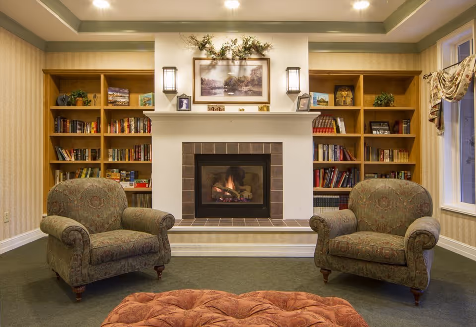 A cozy living room with two patterned armchairs facing a fireplace with a lit fire. The fireplace is flanked by built-in wooden bookshelves filled with books and decorative items. Above the fireplace is a framed landscape painting and floral arrangement, with two wall-mounted lantern-style lights on either side. A large window with floral curtains is visible on the right side.
