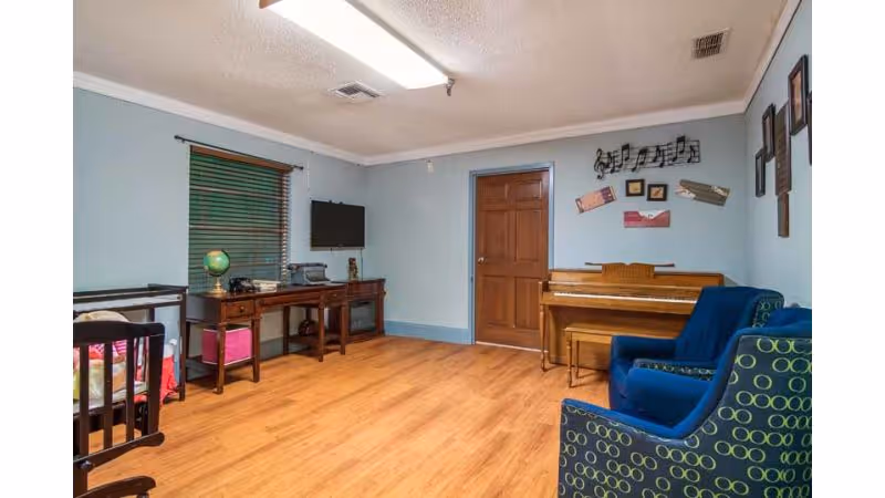 A cozy room with light blue walls and wooden flooring featuring a wooden piano against the far wall, a blue armchair with a circular pattern, a wooden desk with a globe and other items, a wall-mounted TV, and a wooden door. Musical notes and framed pictures decorate the wall above the piano.