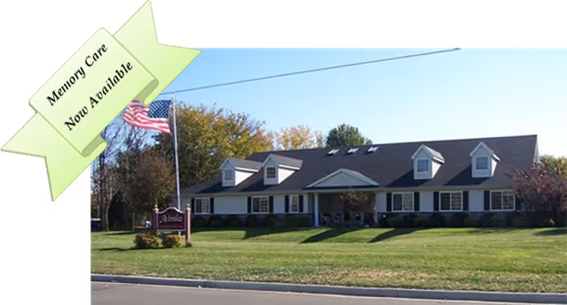 Exterior view of a single-story building with a dark roof and white siding, surrounded by a well-maintained lawn and trees. An American flag is flying on a flagpole near a sign that reads Avonlea Cottage. A banner in the top left corner states Memory Care Now Available.