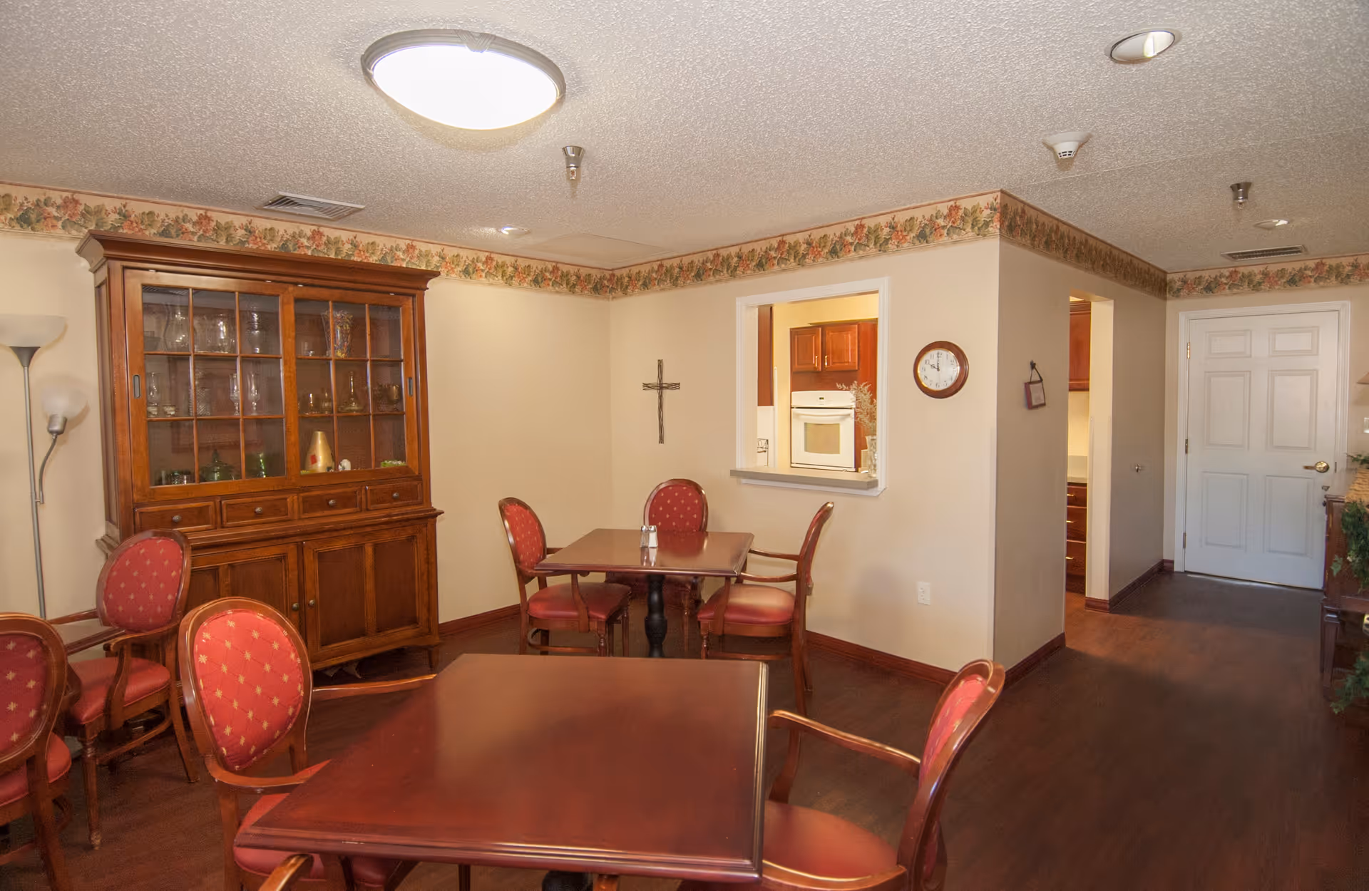 A cozy dining area in an assisted living community featuring wooden tables and red cushioned chairs. There is a wooden cabinet with glass doors displaying glassware, a wall clock, a cross on the wall, and a pass-through window to the kitchen. The room has floral wallpaper border near the ceiling and hardwood flooring.