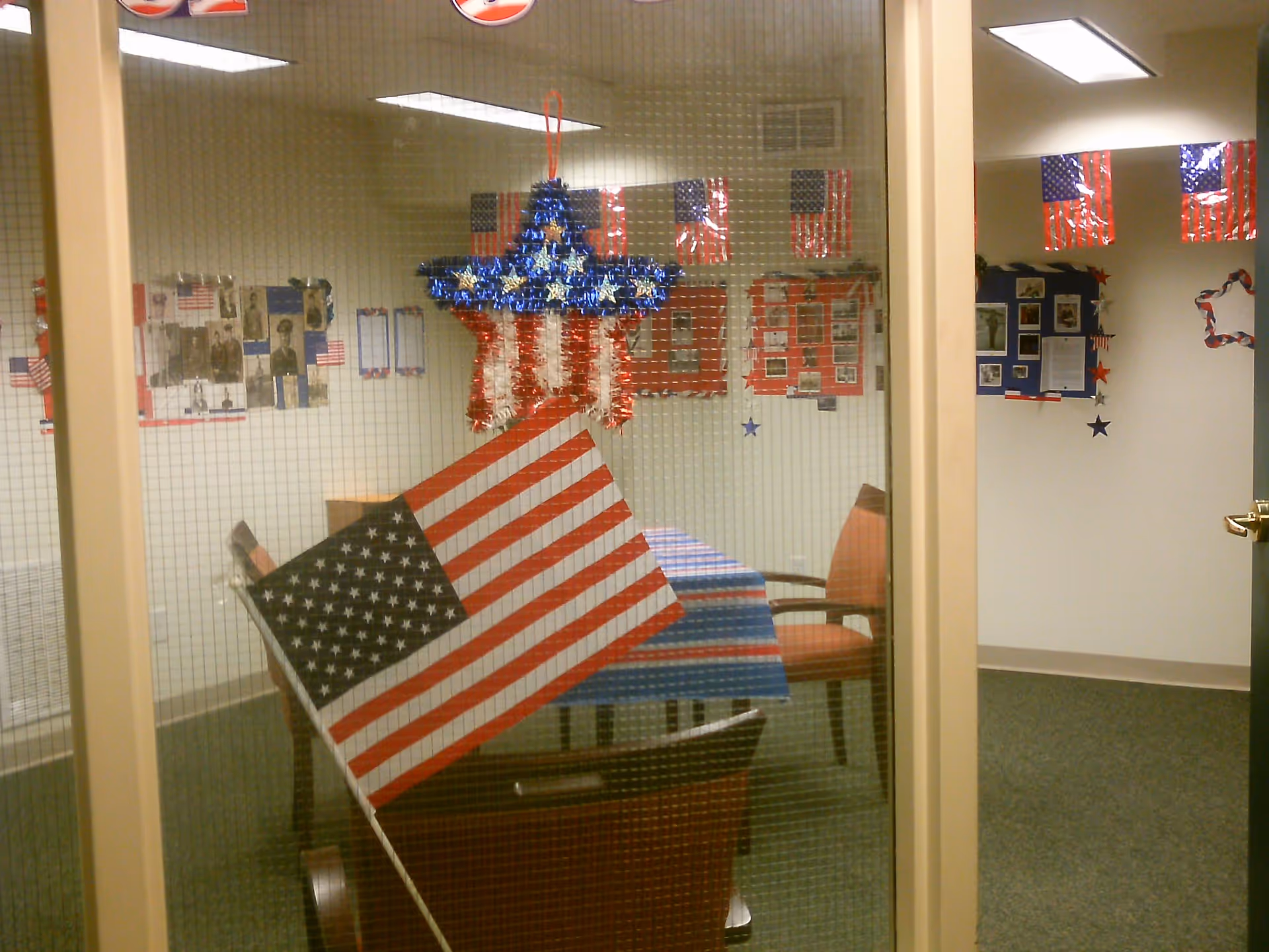 View through a glass door with wire mesh showing a room decorated with American flags and patriotic decorations. Inside, there is a table covered with a red, white, and blue striped tablecloth, surrounded by chairs. The walls are adorned with various patriotic posters and pictures.