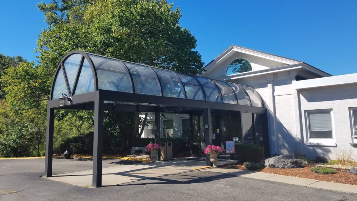 Entrance of Cedar View Rehabilitation & Healthcare Center featuring a covered walkway with a curved transparent roof supported by black metal frames. The building exterior is white with large windows, surrounded by greenery and potted flowers near the entrance.