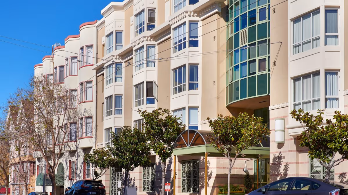 Exterior view of a multi-story residential building with large windows and a green awning entrance, surrounded by small trees and parked cars under a clear blue sky.