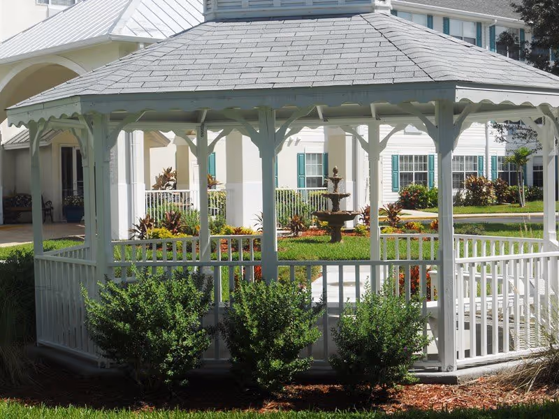 White wooden gazebo with a shingled roof surrounded by green bushes and grass, with a multi-tiered fountain and a white building with green shutters in the background.