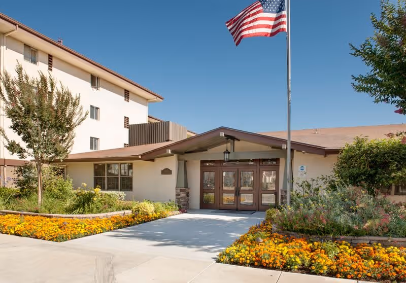 Entrance of a senior living facility named Ararat Gardens with a pathway leading to double wooden doors under a peaked roof. The building is surrounded by well-maintained flower beds with yellow and orange flowers, green shrubs, and trees. An American flag is flying on a flagpole near the entrance.