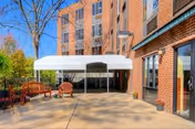 Outdoor patio area at Green Meadows Nursing & Rehabilitation Center with a white canopy, wooden benches, potted plants, and a brick building facade under a clear blue sky.