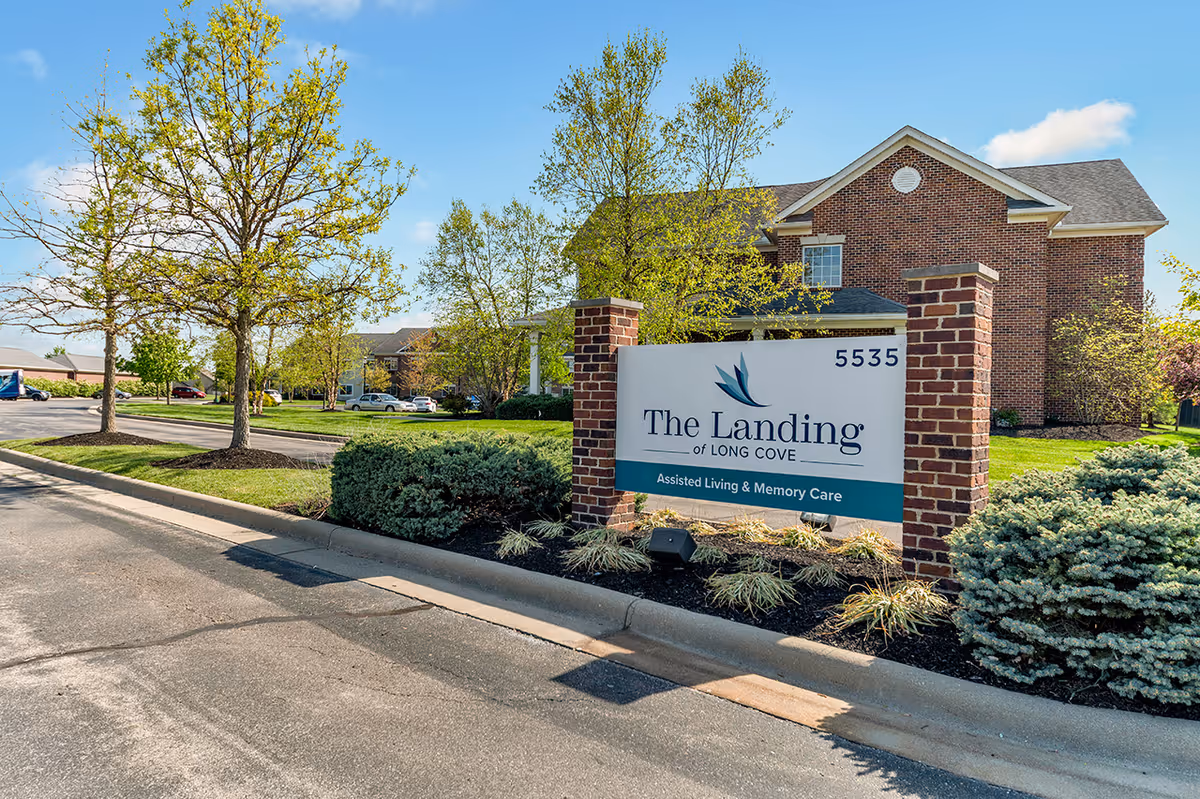Exterior view of The Landing of Long Cove assisted living and memory care facility with a brick building, a sign displaying the facility name and address 5535, surrounded by trees and shrubs under a clear blue sky.