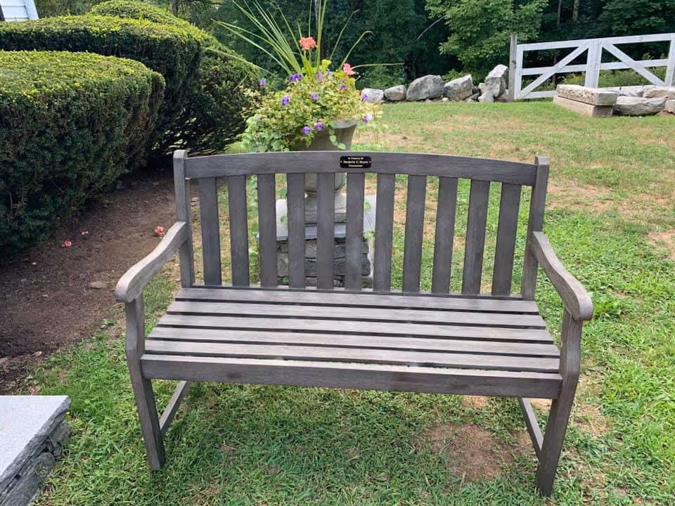 Wooden outdoor bench on grass with a planter, trimmed shrubs, and a white fence in the background.