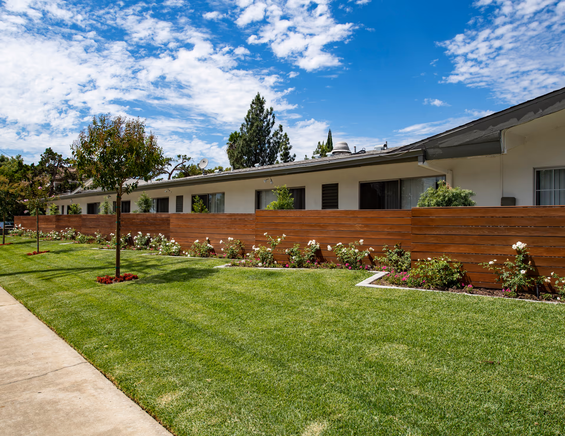 Green lawn, flowerbeds and a wooden privacy fence in front of a single-story care facility building under a blue sky.