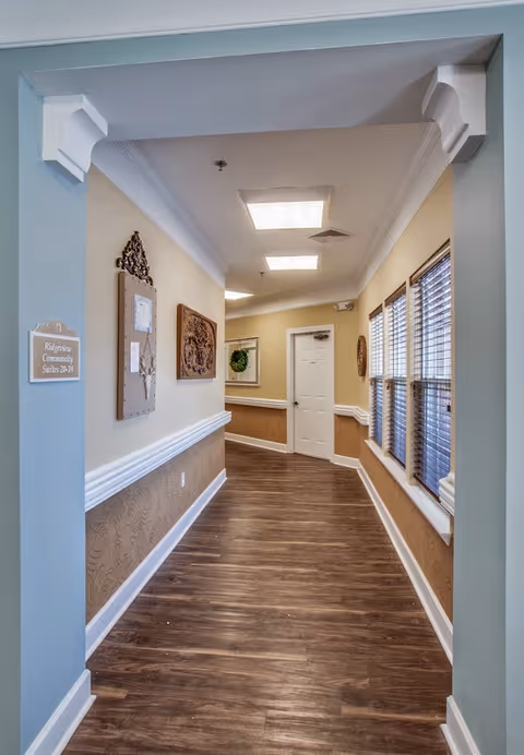 A well-lit hallway in a senior living facility with wood flooring, beige and white walls, decorative wall hangings, and windows with blinds on the right side. A white door is visible at the end of the hallway.