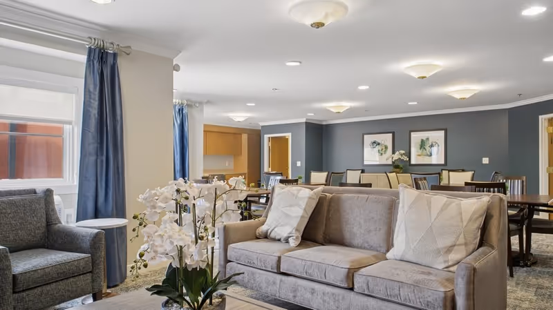 Communal senior living room with a sofa and armchairs in the foreground, dining tables in the background, and decorative orchids.