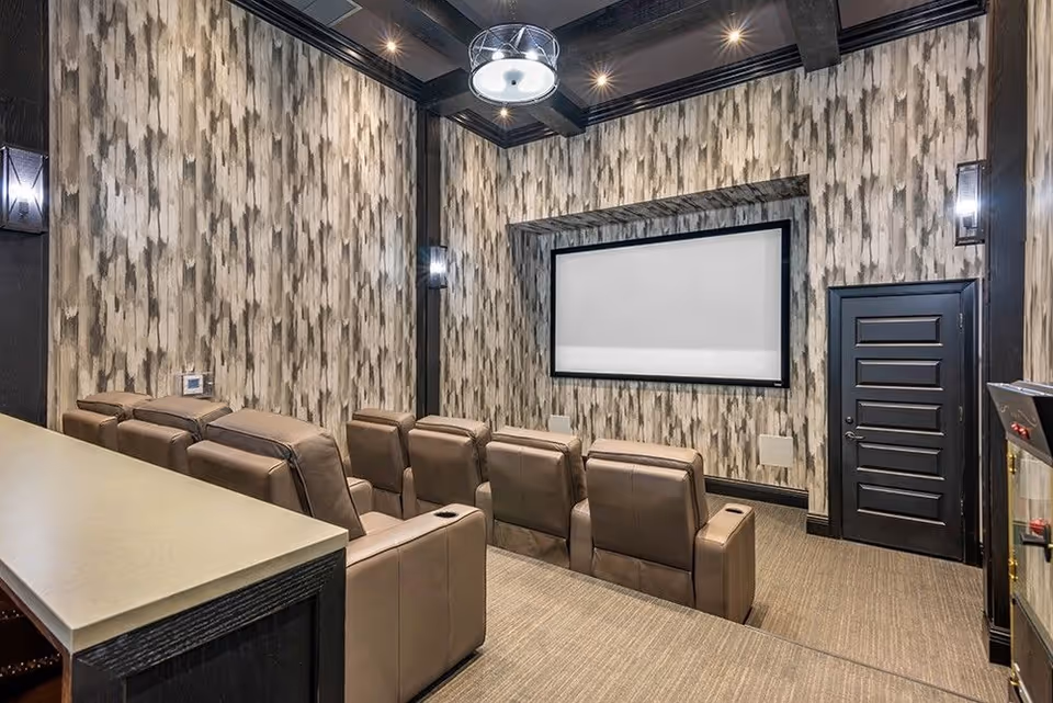 A small home theater room with two rows of brown leather recliner chairs facing a large blank projection screen. The walls are decorated with patterned wallpaper in shades of gray and beige, and the ceiling has dark wooden beams with recessed lighting and a central modern light fixture. There is a black door to the right and a counter or bar area to the left.