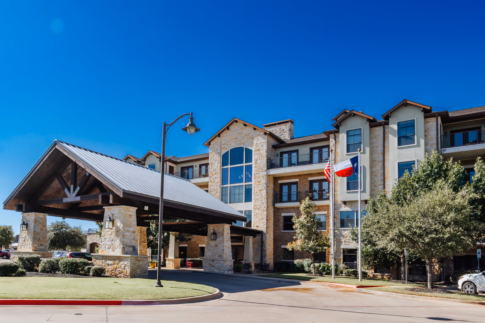 Exterior view of Waterview The Point Independent Living facility showing a multi-story building with stone and brick facade, a covered entrance with wooden beams and a metal roof, two flagpoles with the American and Texas flags, trees, and a clear blue sky.