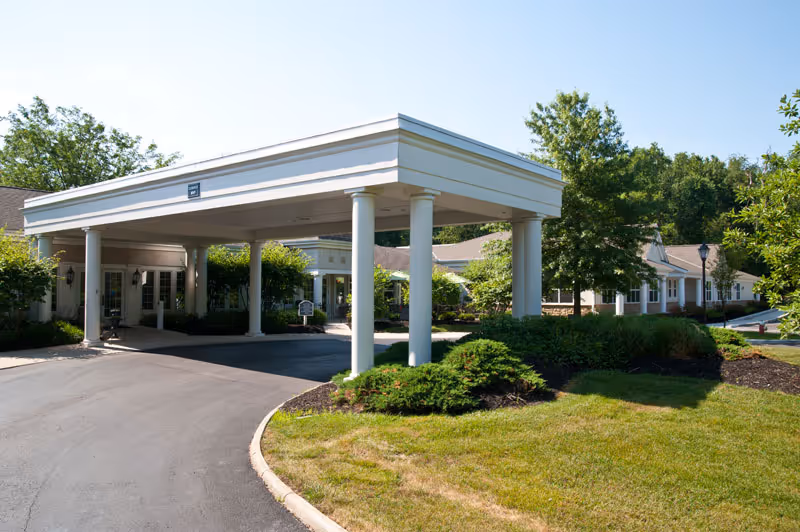 Front entrance of a senior living facility with a covered porte-cochère, white columns, driveway and landscaped grounds.