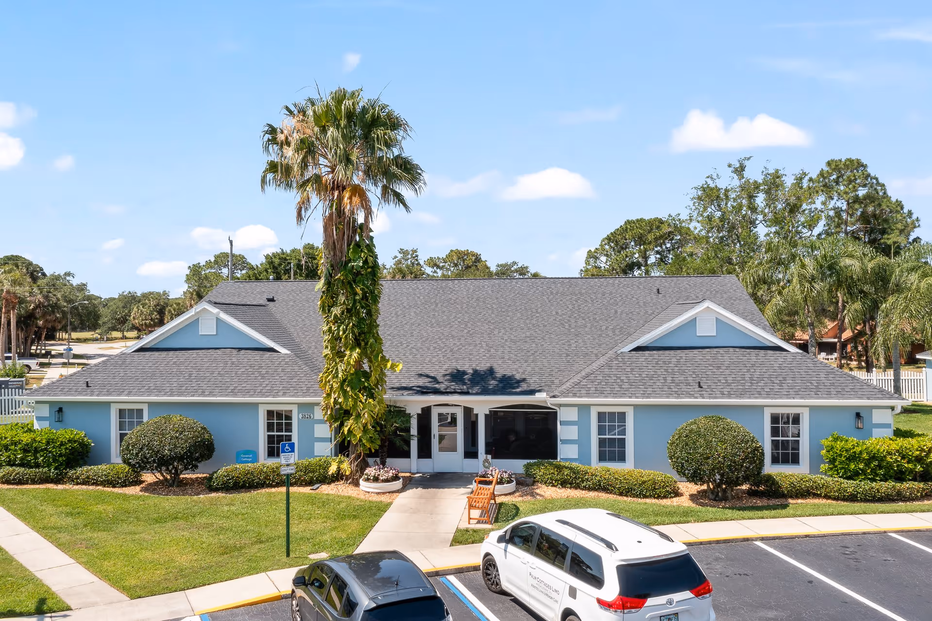 Exterior view of a single-story blue building with a gray roof, surrounded by neatly trimmed bushes and a tall palm tree in front. There is a sidewalk leading to the entrance with a bench nearby, and two parked cars in front of the building under a clear blue sky.