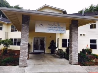 Entrance of Suzanne Elise Assisted Living Community featuring a covered walkway supported by brick pillars, with a sign above the entrance displaying the facility's name and a message about opening soon. The building is light-colored with multiple windows and surrounded by landscaped greenery.