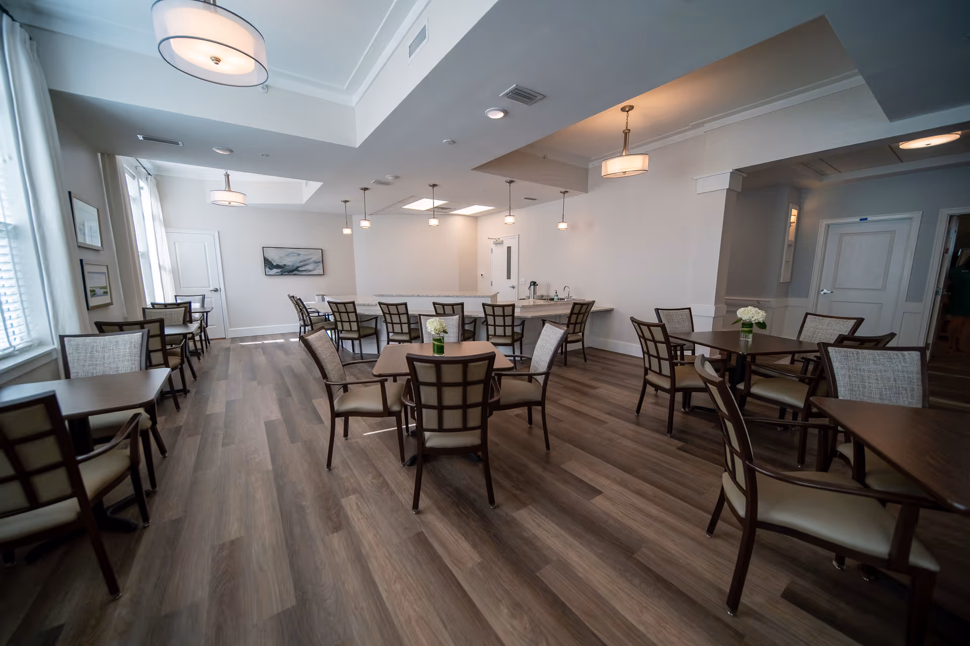 A bright and spacious dining room with multiple square wooden tables and cushioned chairs arranged neatly. The room features large windows with white blinds allowing natural light to fill the space. Modern pendant lights hang from the ceiling, and a countertop with additional chairs is visible at the far end. The walls are painted white with minimal artwork, and the floor is covered with wood-style flooring.