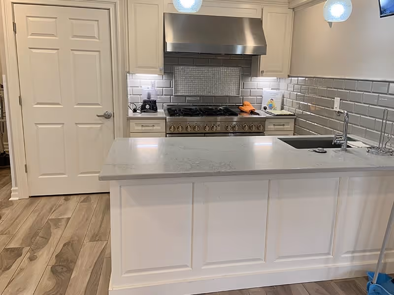Modern kitchen with white cabinetry, a large island with a gray countertop, a stainless steel stove and range hood, subway tile backsplash, and a sink with a faucet. The floor has wood-like tiles and there is a closed white door on the left side.